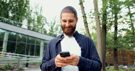 Smiling bearded Caucasian man typing text messages on his smartphone while walking along an urban street. Concept of mobile communication, technology, modern lifestyle, business, IT, startup