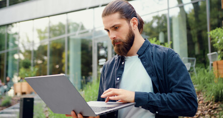 Fototapeta premium Handsome entrepreneur using a wireless laptop outdoors near a modern office building, typing and scrolling while managing online business and remote work.