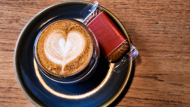 A cup of coffee, latte or cappuccino with latte art that has a heart-shaped design in the milk foam, served with a single Lotus Biscoff cookie. 