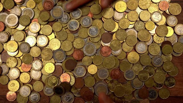 Close-up of a woman's hands moving slowly and caressing a large pile of euro coins on a wooden table. The scene represents wealth and greed