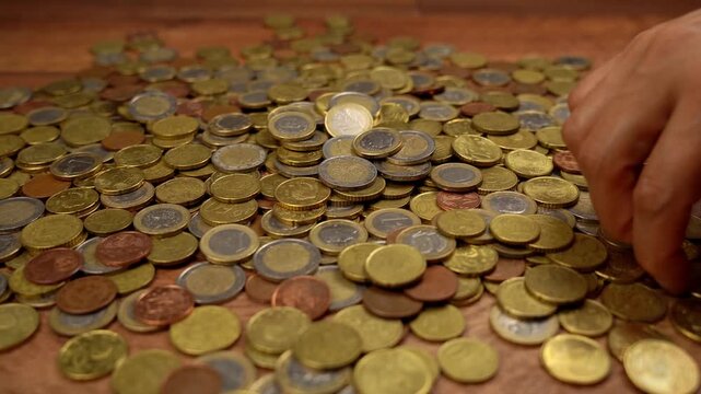 Close-up of a person's hand sorting through a large pile of various euro coins on a wooden table, counting and stacking the money