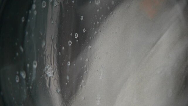 A close-up of dirty white laundry spinning and falling inside the drum of a washing machine. Bubbles of foam run down the glass of the washing machine's door.