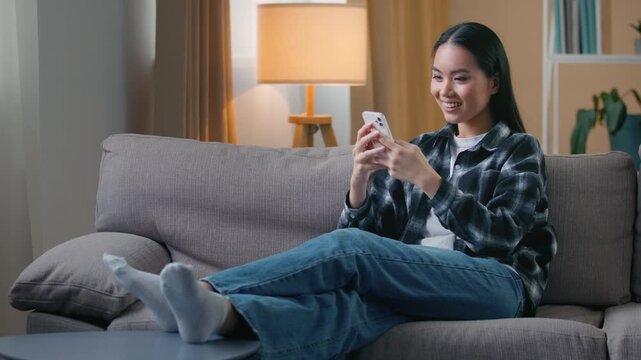 Asian woman sitting sofa at home using smartphone browsing social media relaxing Korean female girl watch news scrolling shopping online choosing clothes dating app wifi internet store leisure indoors