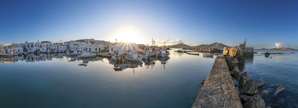 Panorama, fishing boats in the harbour of Naoussa at sunset, reflected in the sea, sun star, ruins of the Venetian castle on the right, white Cycladic houses, Naoussa, Paros, Cyclades, Greece