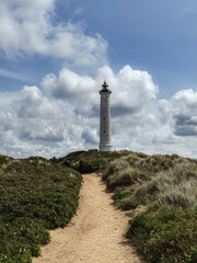 tall coastal lighthouse at end of sandy path winding through grassy dunes and wildflowers. beneath dramatic sky with billowing clouds, guidance, resilience, and seaside tranquility.