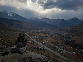 Scenic mountain landscape featuring a stone cairn in the foreground, overlooking a valley with a winding road and distant snow-capped peaks under a dramatic cloudy sky