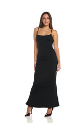 Studio shot of a smiling woman with dark hair wearing a long black dress and high heels, standing in a relaxed pose, full body, on a white background
