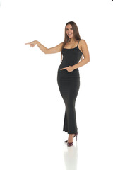 Studio shot of a smiling woman with dark hair wearing a long black dress and high heels, standing and pointing to the side and down, full body, on a white background