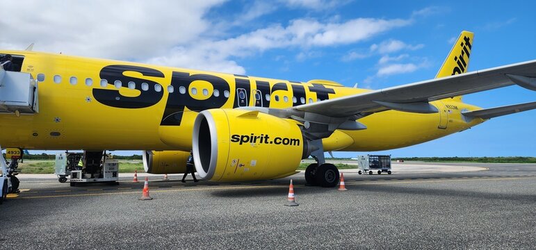A bright yellow Spirit Airlines passenger jet is parked on the airport tarmac during ground operations, captured in a clean side profile under a partly cloudy blue sky. 
