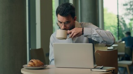 Concentrated Indian businessman with laptop working drinking tea coffee with croissant in cafe Arabian business man with computer job focused drink serious professional work online internet guy male