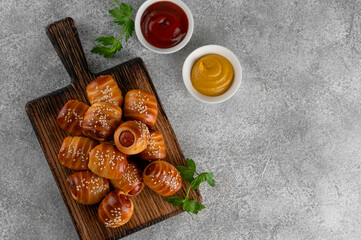 Mini hot dog bites with mustard and ketchup. Sausages baked in a yeast dough cover. Pigs in a blanket on a wooden board on a gray background. Top view, copy space.