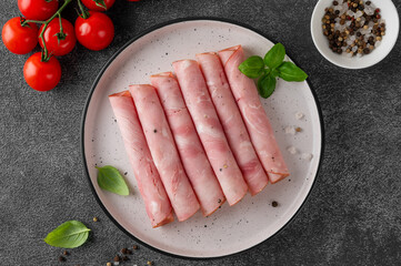 Delicious ham cut into round slices on a plate with spices, cherry tomatoes and basil leaves on a dark stone background. Top view.