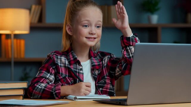 Caucasian kid daughter girl writing online studying raising hand ready to answer online teacher smiling clever school child pupil learning distant from home laptop video call e-learning write notes