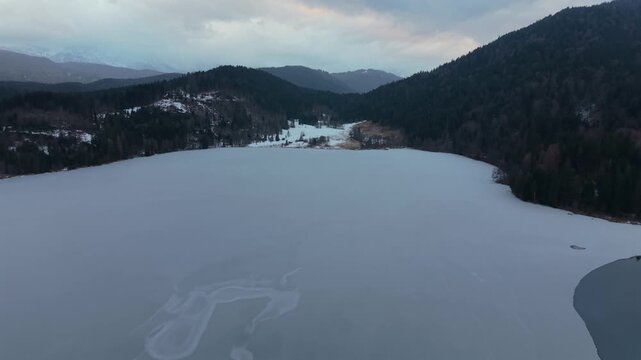 Eco tourism spot in winter. Drone footage of a pristine alpine lake. Cinematic aerial view of Lake Barmsee, Bavaria. Winter drone footage reveals frozen surface, forest and Karwendel mountains.