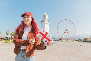 Smiling woman with pink hair standing outdoors, holding Georgian flag with Batumi lighthouse and Ferris wheel in background