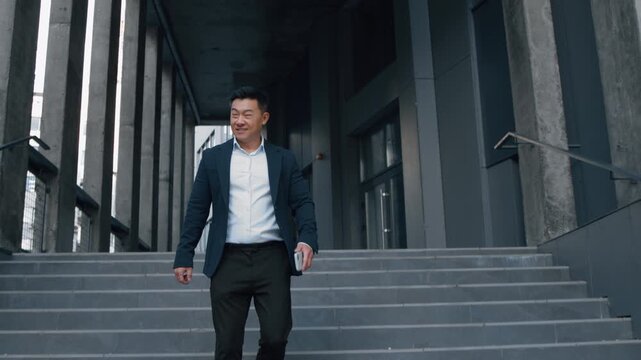 Happy smiling Asian business man walking down stairs in city Korean businessman leaving office company building move go walk going Chinese Japanese middle-aged male with phone professional manager
