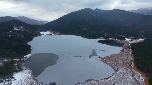 Aerial drone footage of Barmsee in Kruen, Oberbayern. Winter scenery with frozen water, forest and Karwendel mountain range in the background. Luftaufnahme per Drohne vom Barmsee in Bayern