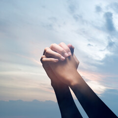 Closeup image of hands clasped together in prayer against a backdrop of a peaceful and soft colored sky. Concept of faith, hope, and spirituality
