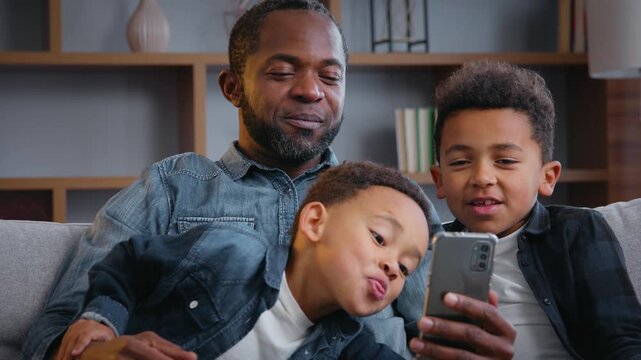 African American happy family with mobile phone smiling taking picture selfie grimacing father two kids sons boys children looking in smartphone trying funny photo mask recording video call at home