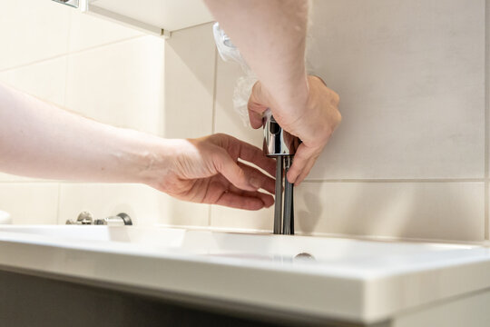 Person installing a chrome bathroom faucet on a white sink. Hands thread the base while holding the top steady in a modern tiled bathroom during sink installation or renovation