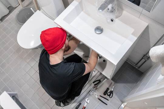 Handyman installing a new faucet and plumbing lines under a bathroom sink. Tools on a tiled floor in a modern bathroom illustrate home renovation, repair, maintenance, and proper installation work