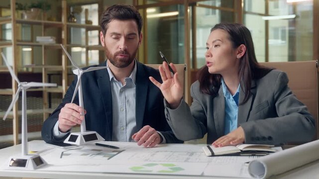 Korean environmental analyst female consultant Caucasian man project conversation sustainable energy holding hands windmill model eco blueprint multiethnic group power solution innovation discussion