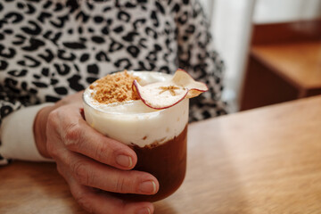A close-up of a person's hand holding a glass of iced coffee. The beverage is topped with a thick layer of white foam, brown crumble topping, and a thin slice of dried apple.