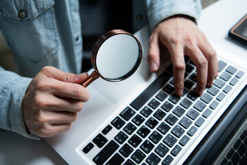 Hand holding magnifying glass over laptop keyboard. Professional freelancer searching for information. Concept of data analysis, security audit, web research, investigation and finding solutions.