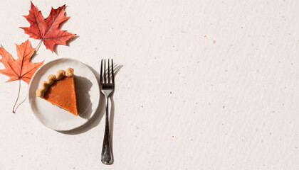 Slice of pumpkin pie on white plate with fork beside it, accompanied by two autumn leaves on textured background, evokes cozy Thanksgiving atmosphere