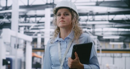 Woman in Safety Helmet Conducts Audit, Swiping Screen of Handheld Tablet to View Production Metrics. Backdrop is Automated in Modern Industry 4.0 Plant. Data Analysis and Digital Transformation.