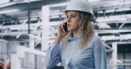 Fototapeta premium Female Engineer Wearing Hard Hat Walks Across Production Workshop and Speaks on Mobile Phone, Supervising Processes in High Tech Factory Environment. Modern Industry, Leadership and Workflow Control.