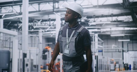 African American Male Factory Supervisor Walking Across High Tech Manufacturing Facility, Using Tablet Check Production Data, Assess Machinery Performance and Oversee Automated Industrial Operations.