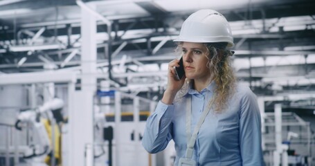 Blonde Curly haired Engineer in White Safety Helmet Walks Through Factory Floor While Talking on Smartphone, Coordinating Operations in Automated Facility. Concept of Communication and Management.