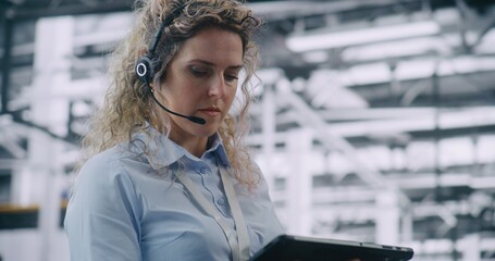 Close Up of Female Factory Engineer Wearing Communication Headset and Actively Engaging With Digital Tablet. Real Time Coordination, Remote Operations, and Digital Communication on Industrial Floor.