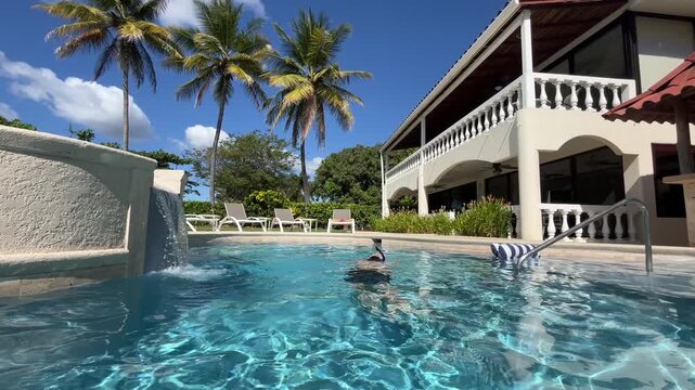 Low tracking shot underwater swimmer at family vacation rental villa with swimming pool and water fall overlooking palm trees at a Tropical destination.