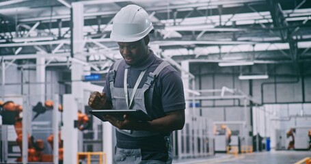 Industrial Male Worker Operating Remote Control System While Overseeing Robots in Fully Automated Production Hall. Concept of Advanced Manufacturing, Smart Industry, Modern Factory Environment.
