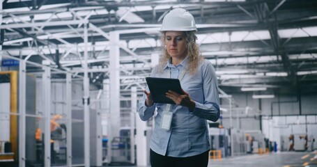 Professional Female Engineer in Hard Hat Checks Production Data on Tablet While Walking Through Modern Automated Factory. Concept of Smart Manufacturing, Industry 4.0, Digital Control and Efficiency.