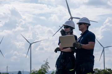 Workers discuss plans while inspecting wind turbines at a renewable energy site during the day with clouds in the sky