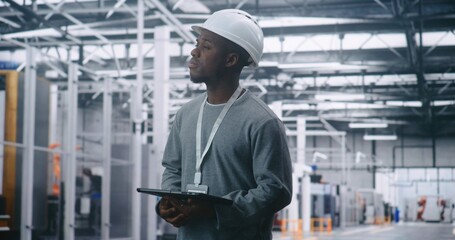 Adult Male Engineer Wearing Safety Helmet, Holding Tablet, and Looking Away to Survey Production Floor. Vast Industrial Interior Emphasizes Strategic Management and Critical On Site Planning.