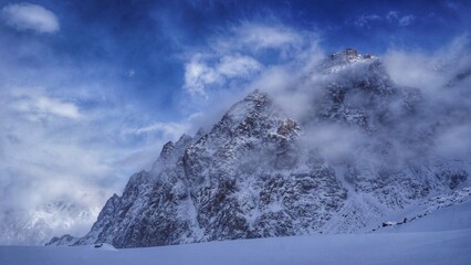 snow covered mountains. A snow-capped mountain peak, partially hidden by fog and clouds, against a cold blue sky.