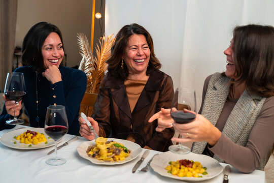 Three women laughing together while enjoying Italian dinner and red wine at home