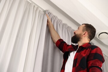 Man hanging up window curtains at home, low angle view. Space for text