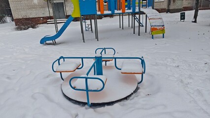 Snow covered playground carousel standing empty in winter yard with slide and play equipment. This image represents childhood pause seasonal contrast and quiet urban playground atmosphere