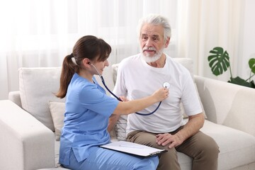 Obraz premium Care worker checking senior man's heartbeat with stethoscope on sofa at home