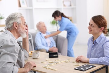 Obraz premium Woman playing dominoes with senior man at table and care worker helping her patient at retirement home, selective focus