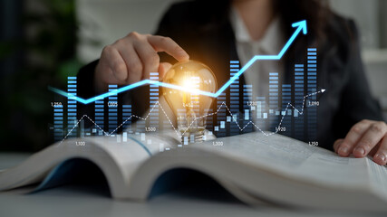 A business professional examines market trends using a light bulb and chart visuals to signify financial growth and innovative strategies in a modern workspace. Scalp