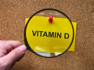 A person with a magnifying glass in his hand magnifying a yellow piece of paper with the words Vitamin D on it. Close up.