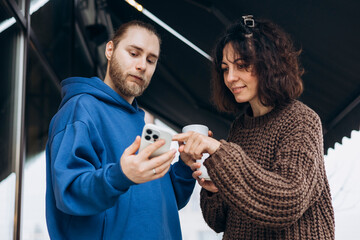 Young man and woman drinking coffee outdoors while looking at smartphone.