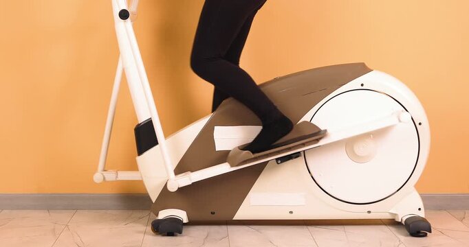 Close-up view of legs exercising on an elliptical trainer at home in a modern interior. This 4K stock video focuses on lower body movement, cardio workout, and indoor fitness activity. Ideal for conce