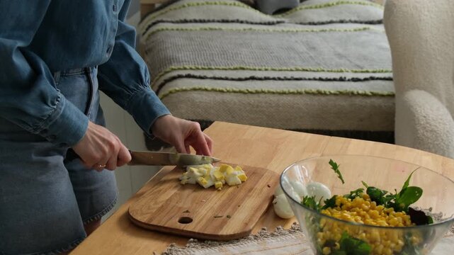 Unrecognizable woman cutting boiled eggs on a wooden board and adding them to a fresh salad with corn and greens in a glass bowl, showcasing a healthy homemade meal preparation process